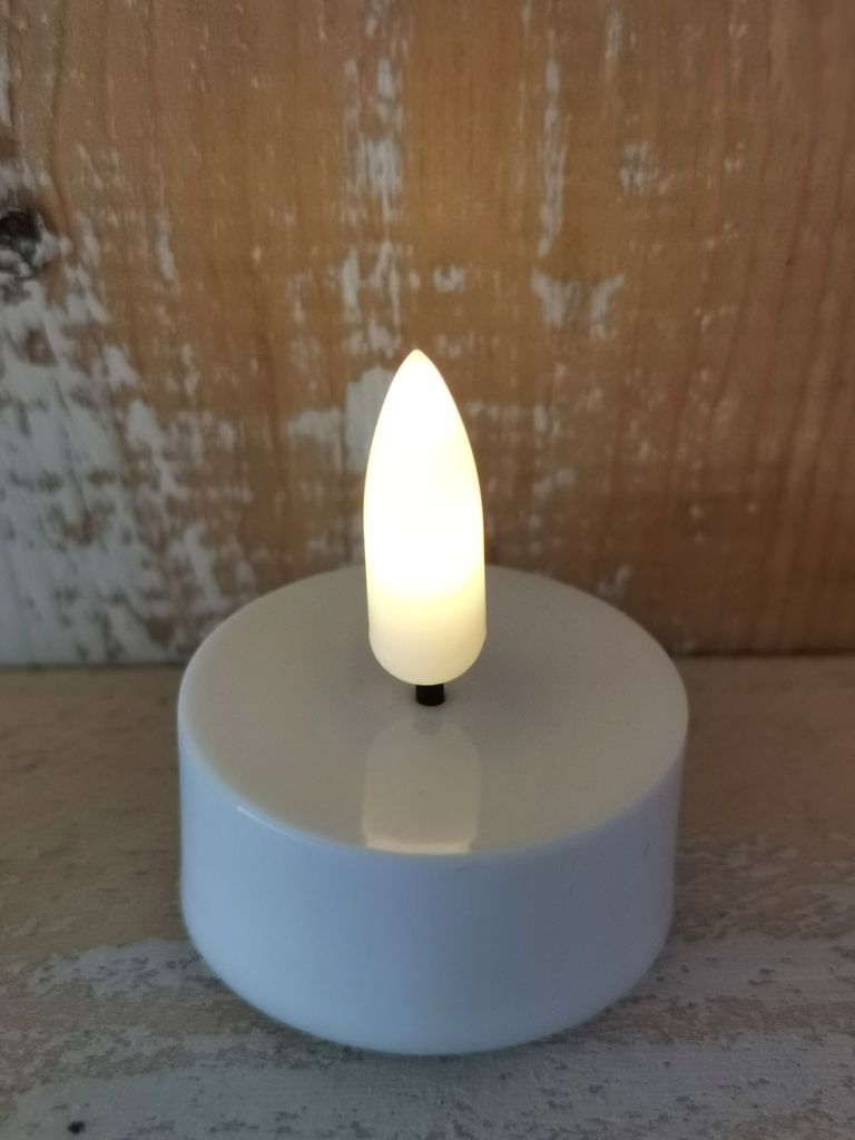 A close-up shot of a white LED tea light with a flickering yellow flame, set against a rustic wooden background.