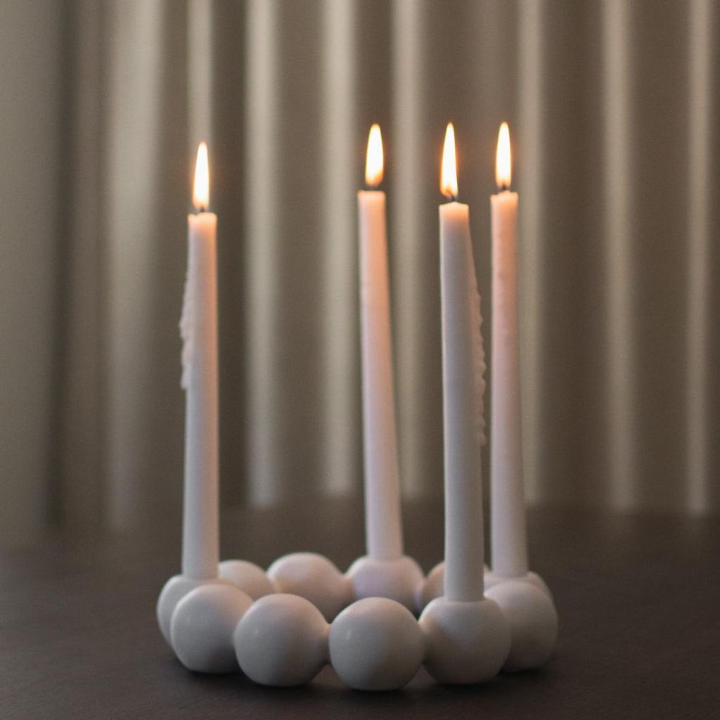 Four white candles are lit and placed in a circular white candle holder on a dark surface, with a textured gray curtain in the background.