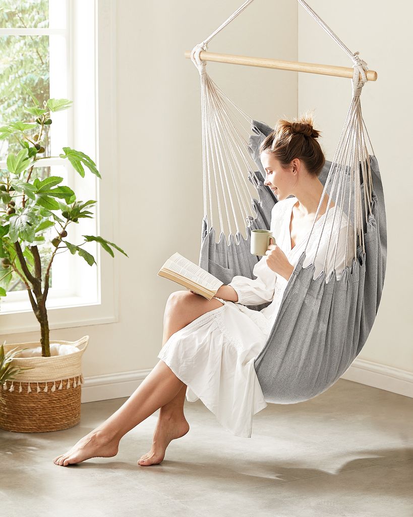 A woman is relaxing in a hanging chair, reading a book and holding a mug.