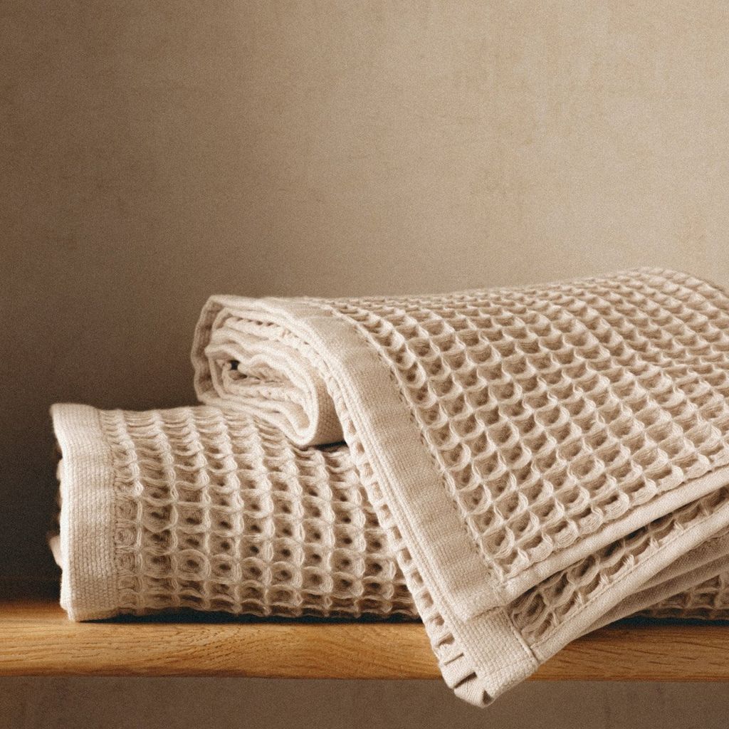 Two folded beige waffle-weave towels are stacked on a wooden shelf against a textured beige wall.