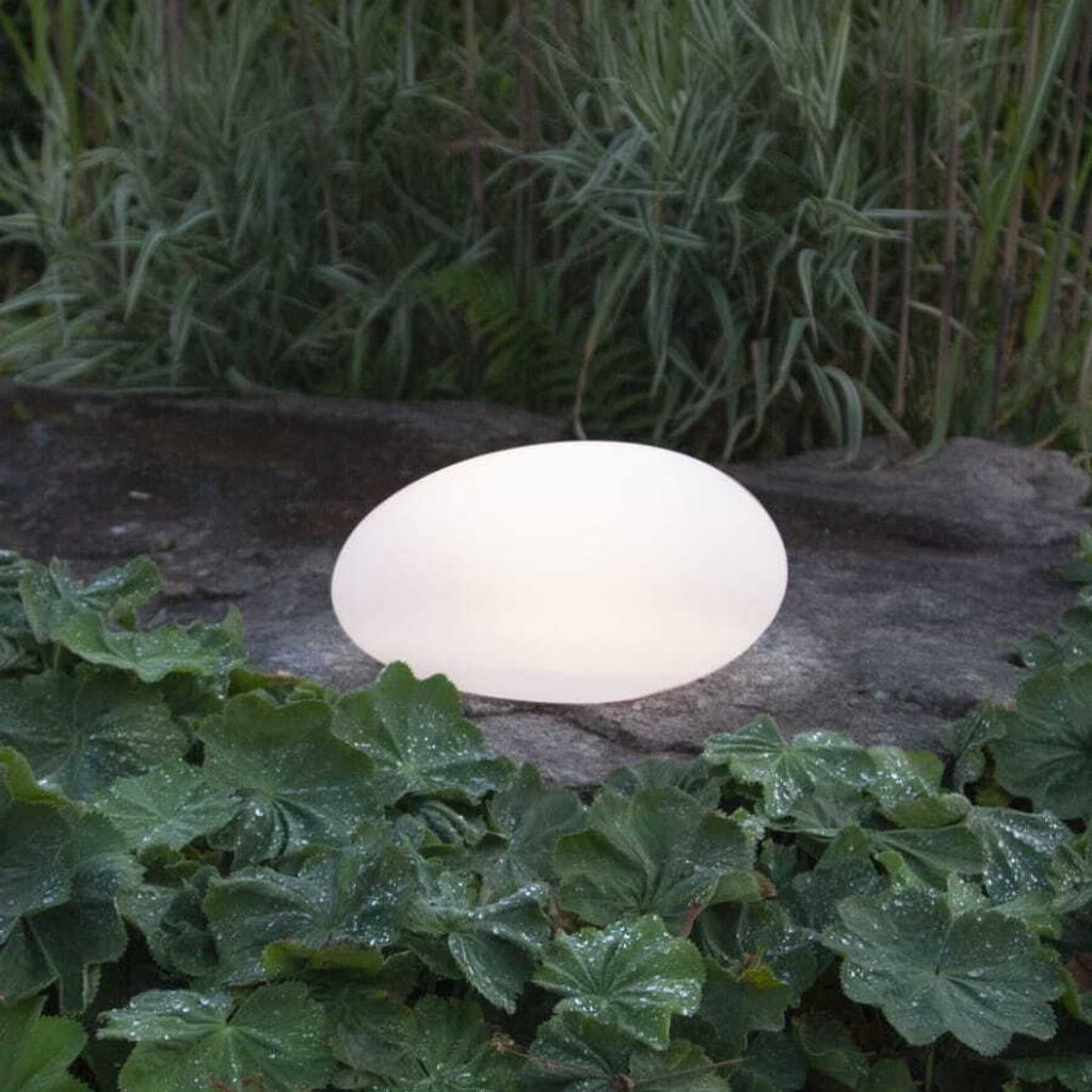 A white, egg-shaped light is placed on a rock in a garden setting, surrounded by green plants.
