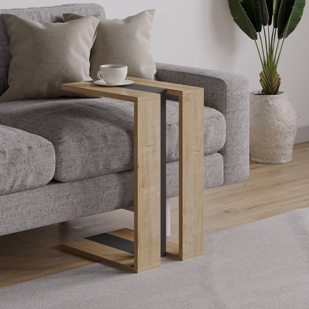 A modern C-shaped side table with a wood finish and dark accents is placed next to a grey sofa with two pillows. A cup of coffee sits on the table. A large potted plant is visible in the background.
