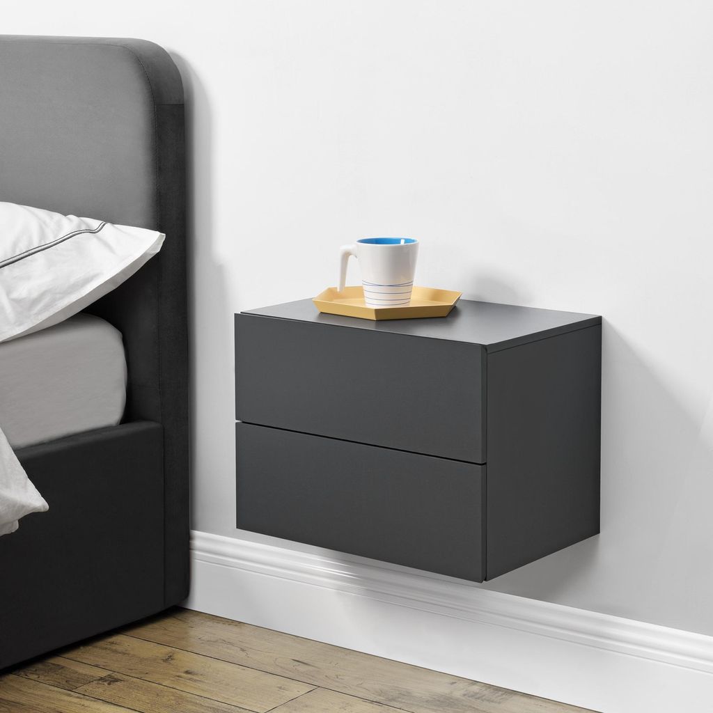 A dark gray floating nightstand with two drawers is mounted on a white wall next to a bed. A white mug with a blue rim sits on a small yellow tray on top of the nightstand.