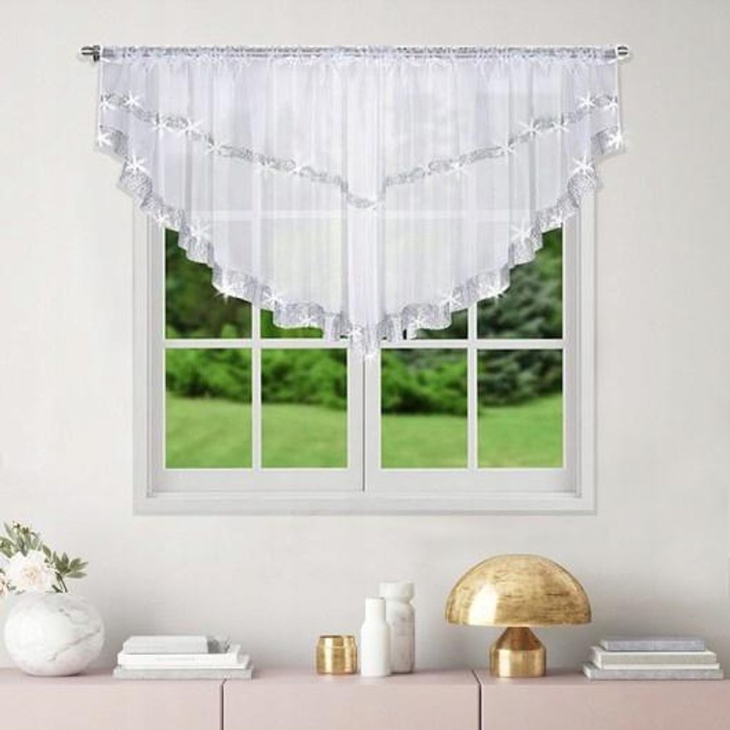 A white valance curtain with silver trim and star-like embellishments hangs over a window with a view of green trees and grass. Below the window, on a pink surface, are decorative items including a vase with flowers, books, a gold lamp, and two white bottl
