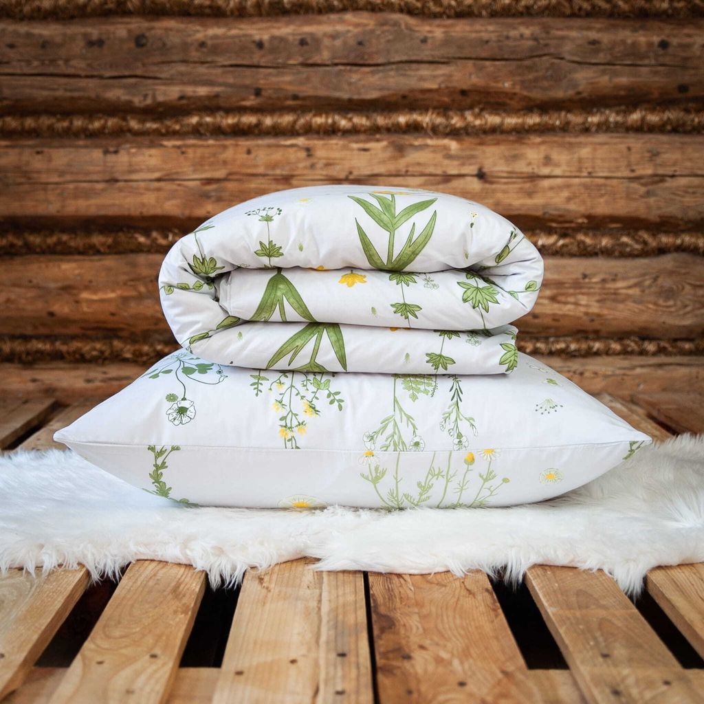A folded duvet and pillow with a floral pattern are stacked on a white fluffy rug, placed on a wooden pallet, with a rustic wooden wall in the background.