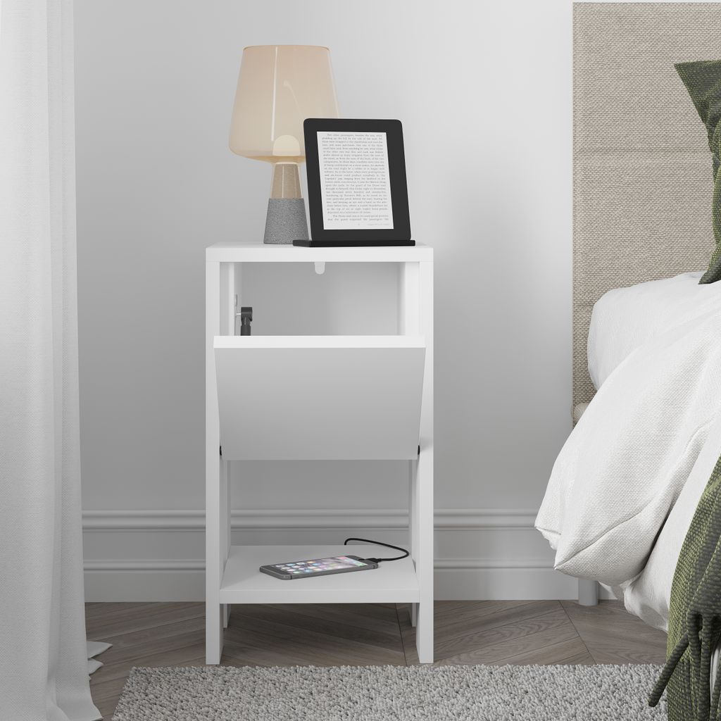 A white nightstand with a lamp and an e-reader on top, and a phone charging on the bottom shelf.