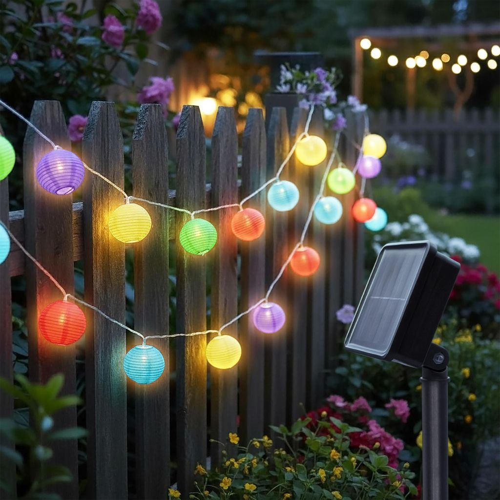 A string of colorful solar-powered lantern lights are strung along a wooden fence in a garden setting.