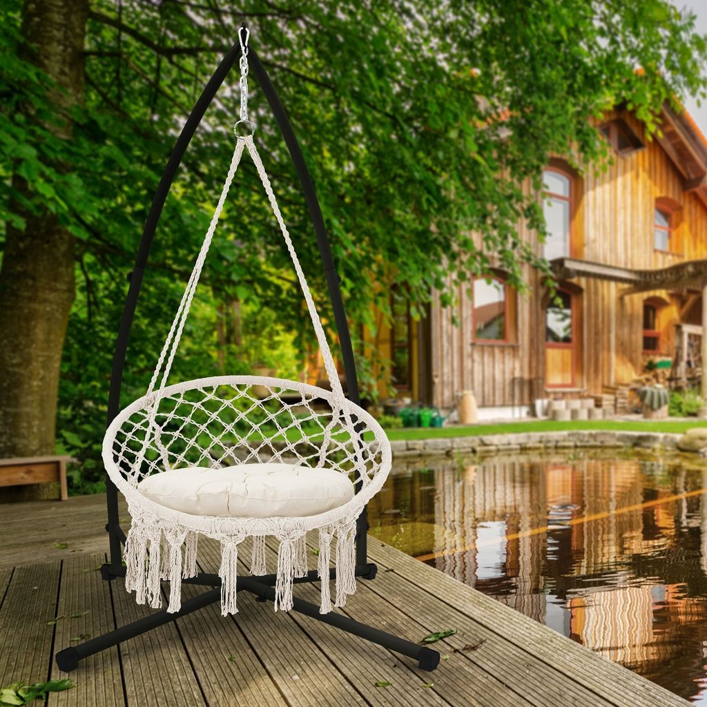 A beige macrame hanging chair with a white cushion is set up on a wooden deck next to a pond, with a rustic wooden house and trees in the background.