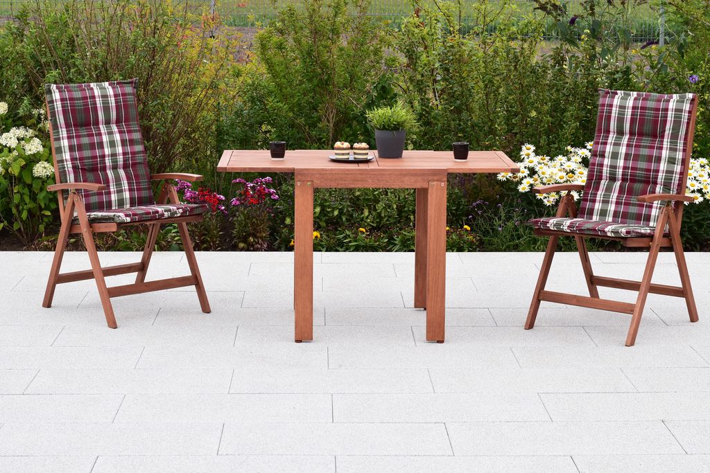 A wooden table and two chairs with cushions are set up on a patio with plants in the background.