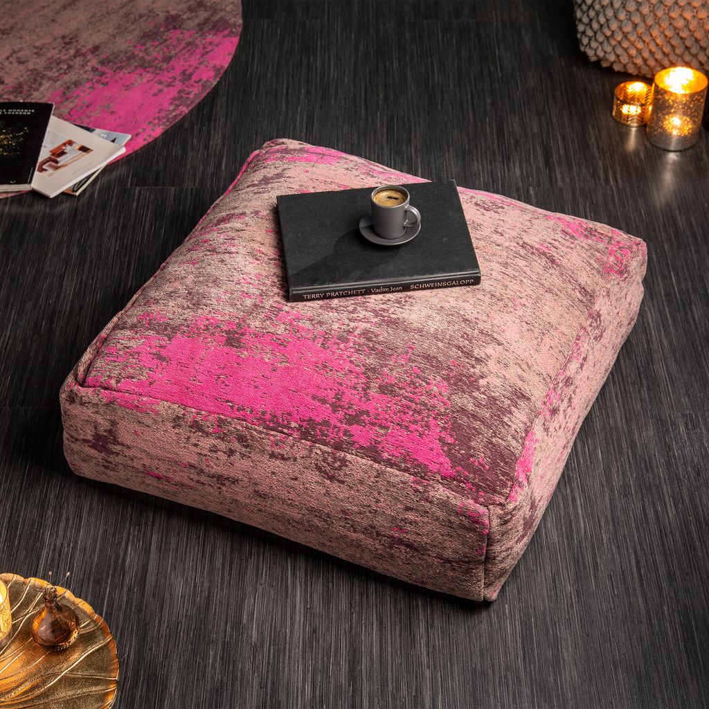 A square floor cushion with a pink and brown abstract pattern sits on a dark wooden floor. A small book and a cup of coffee are placed on top of the cushion. In the background, there are books, candles, and a woven basket.