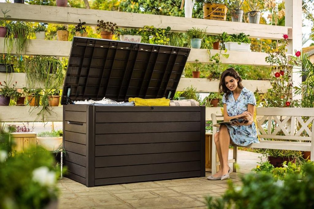 A woman sits on a bench reading a book next to a large storage box on a patio.