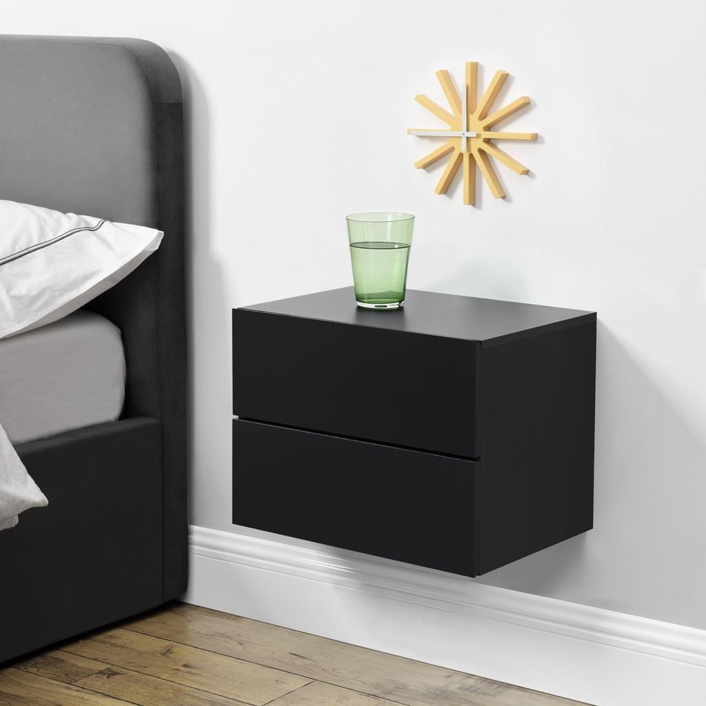 A black floating nightstand with two drawers is mounted on a white wall next to a bed. A glass of water and a wooden clock are on top of the nightstand.