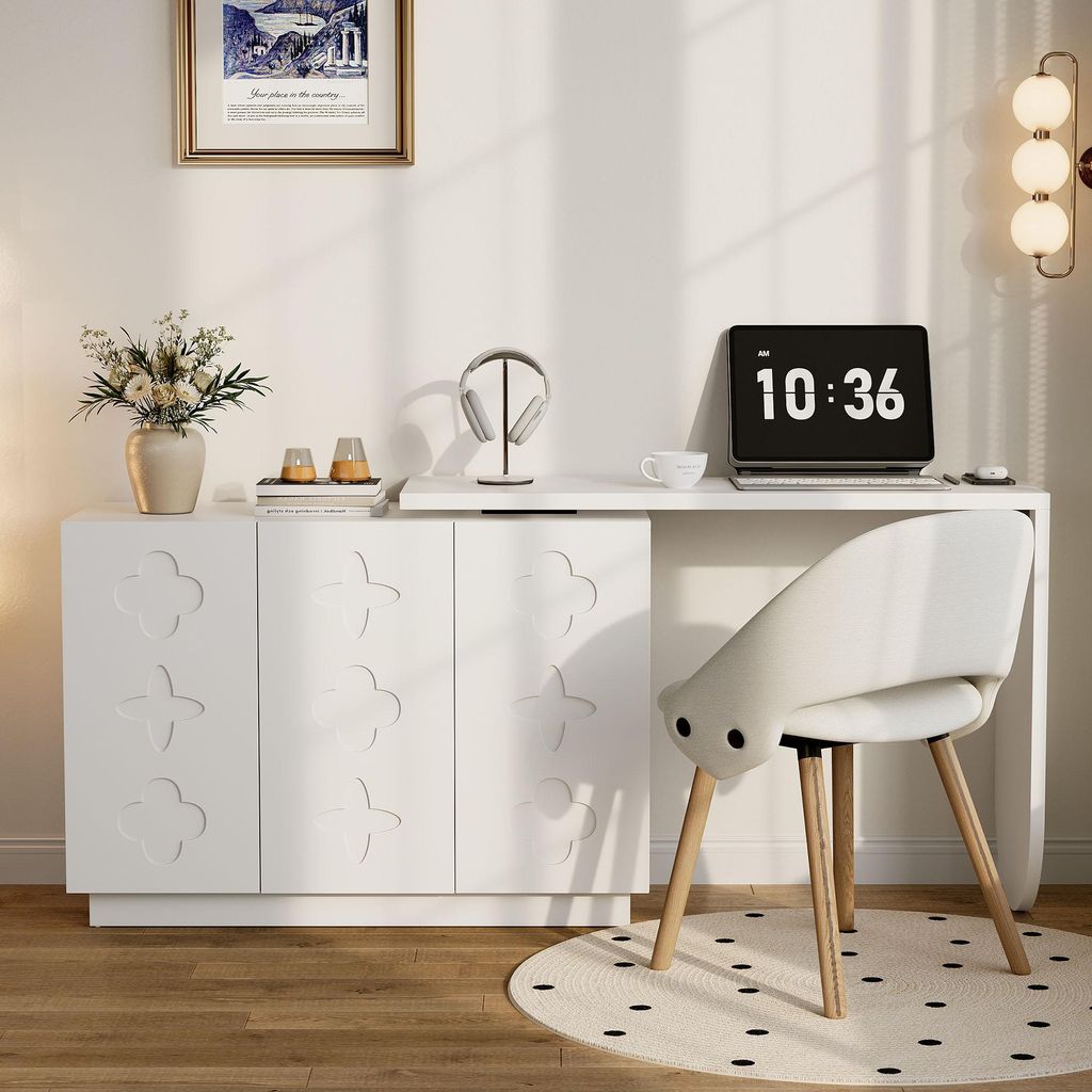 A white desk with a laptop, headphones, and a vase of flowers on top, next to a white cabinet with decorative doors. A white chair is placed in front of the desk.
