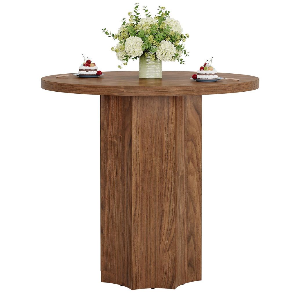 A round wooden dining table with a vase of flowers and two small cakes on top, set against a white background.