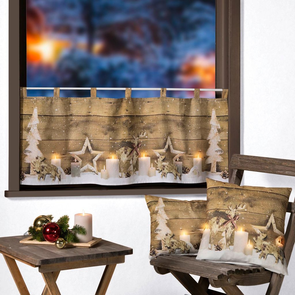 A festive Christmas window decoration with a curtain, candles, stars, and deer, set against a snowy outdoor scene visible through the window. Decorative pillows and a small table with Christmas ornaments are also present.