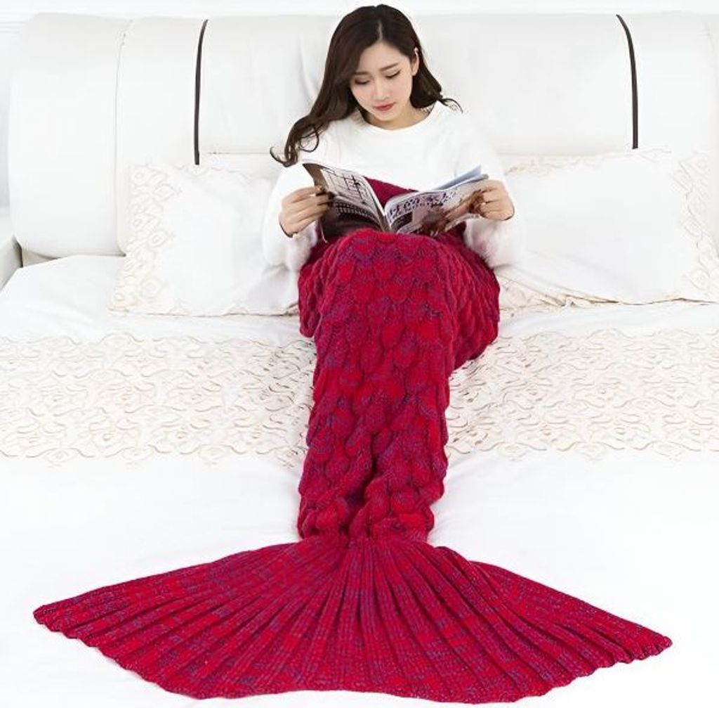 A woman is sitting on a bed, reading a magazine while wrapped in a mermaid tail blanket.