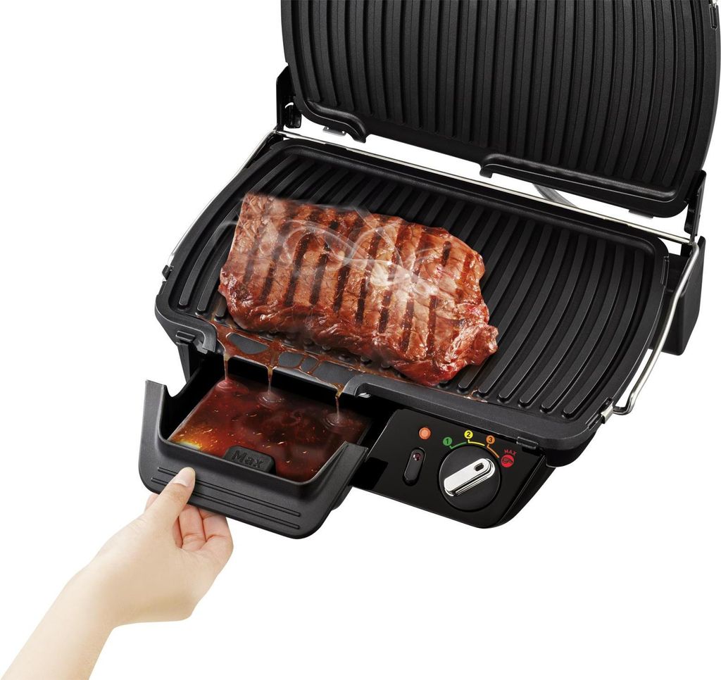 A hand is shown pulling out the drip tray of a black electric grill with a steak cooking on it.
