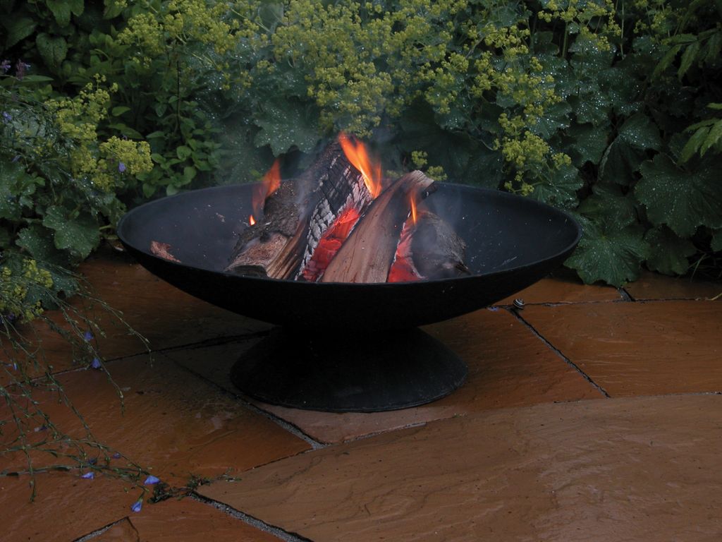 A black fire pit with burning logs sits on a patio with green foliage in the background.