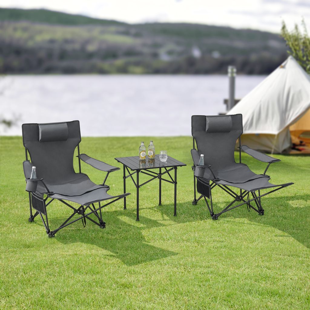Two camping chairs and a small table are set up on a grassy area next to a lake, with a tent in the background.