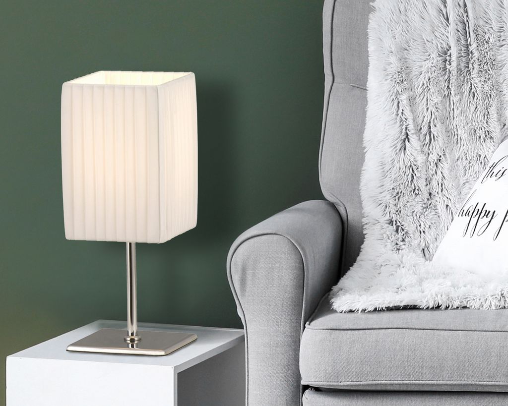 A table lamp sits on a white side table next to a gray armchair with a fluffy gray blanket draped over it.