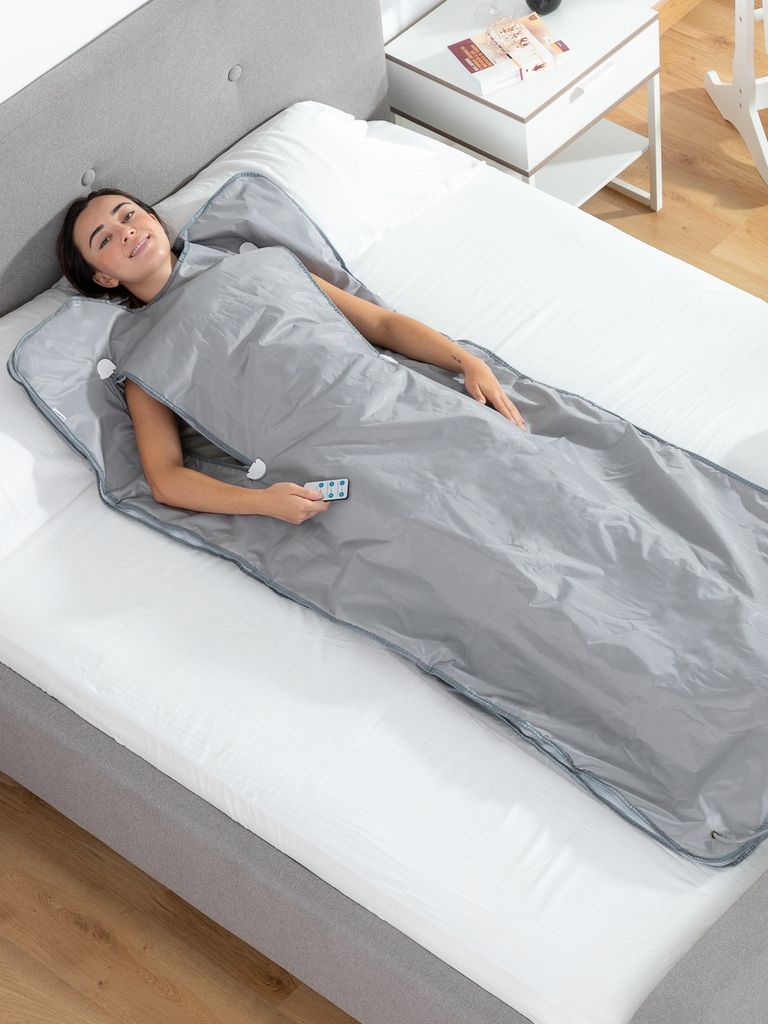 A woman is lying on a bed inside a gray sauna blanket, holding a remote control.