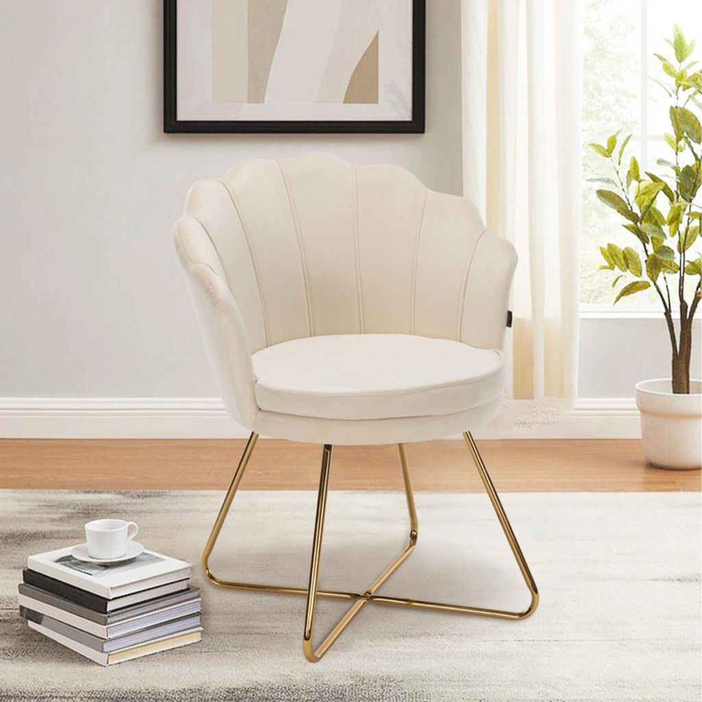 A beige velvet accent chair with gold metal legs is placed on a rug in a room with a framed picture on the wall and a plant in the background.