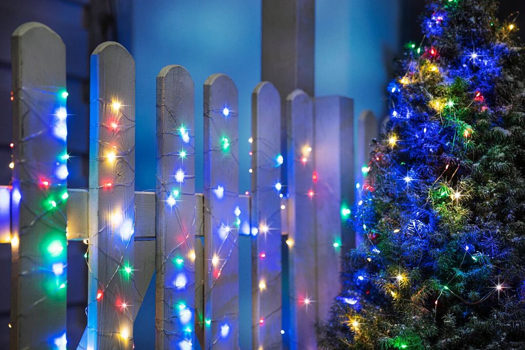 A string of colorful LED lights is wrapped around a wooden fence and a Christmas tree.