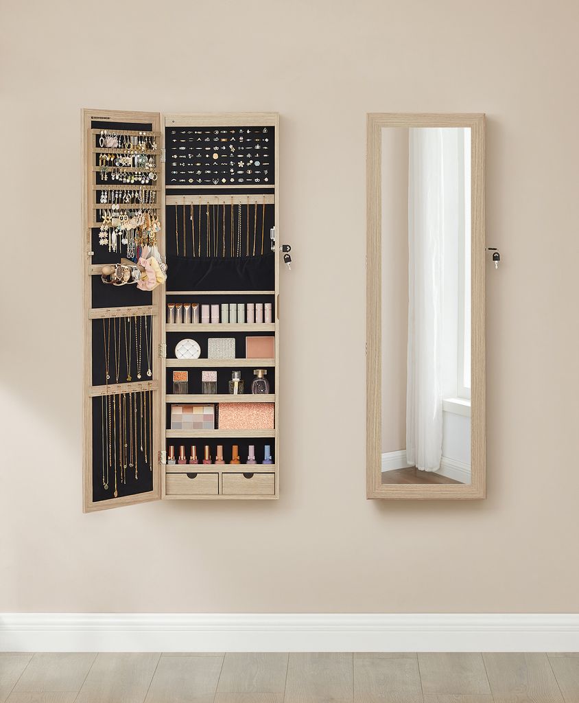 A jewelry cabinet with its door open, displaying various jewelry and makeup items, stands next to a full-length mirror on a wall.
