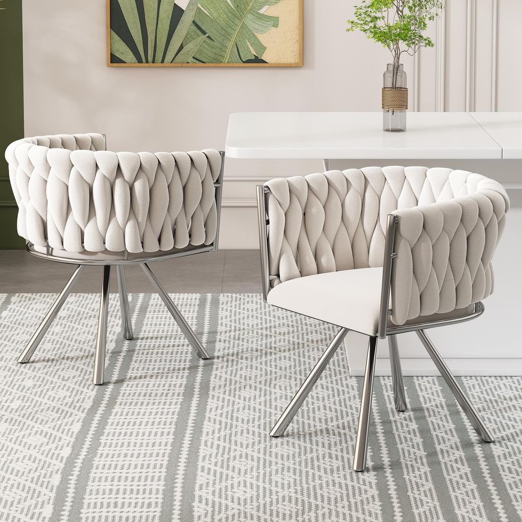 Two beige upholstered dining chairs with silver legs are placed on a grey and white striped rug in front of a white dining table.