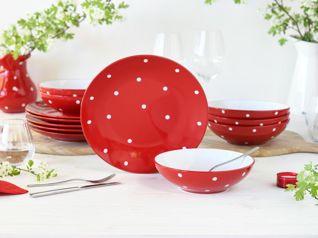 A set of red and white polka dot tableware is arranged on a white surface, with some greenery and flowers in the background.