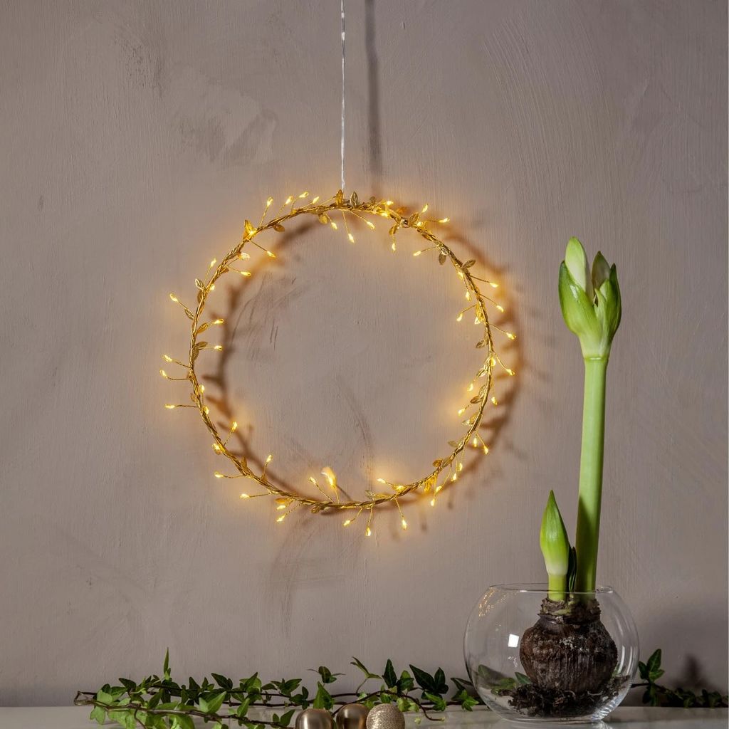 A decorative LED light wreath with warm white lights hangs on a wall, with an amaryllis bulb in a glass vase in the foreground.