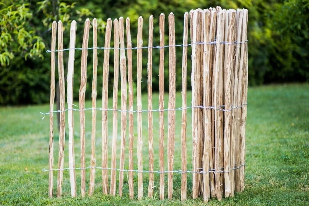 A garden fence made of wooden stakes is shown in a grassy area with trees in the background.