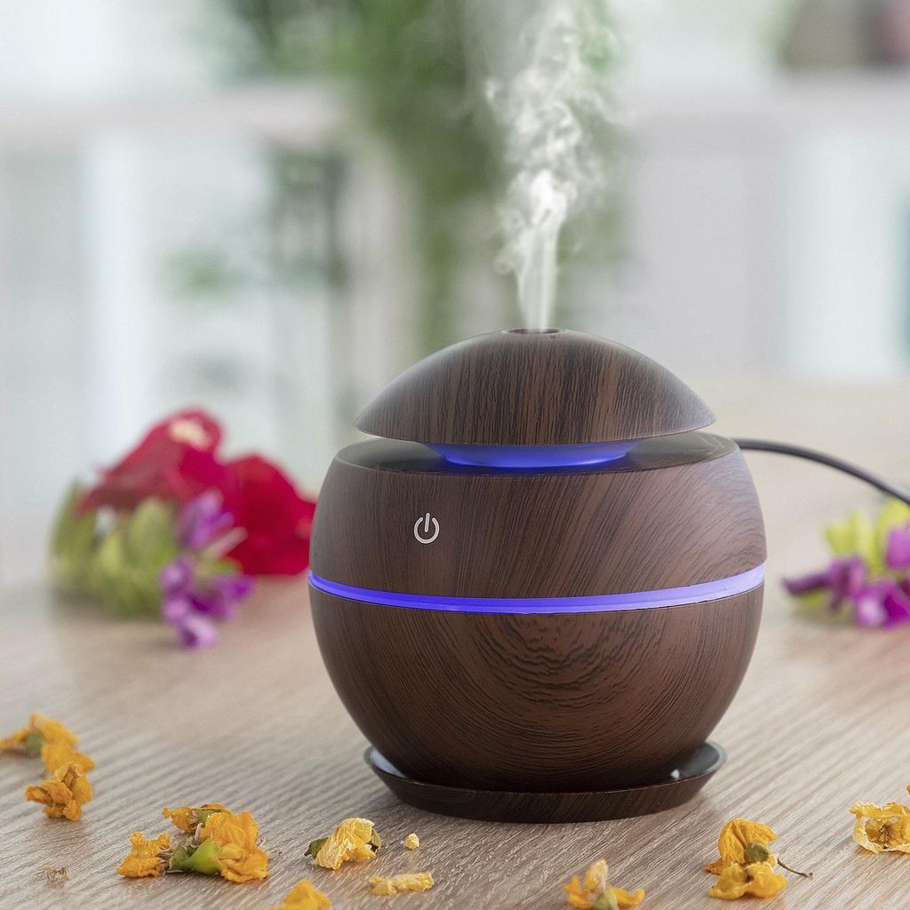 A dark wood-grain mini aroma diffuser emitting a stream of vapor, placed on a wooden table with scattered dried flowers.