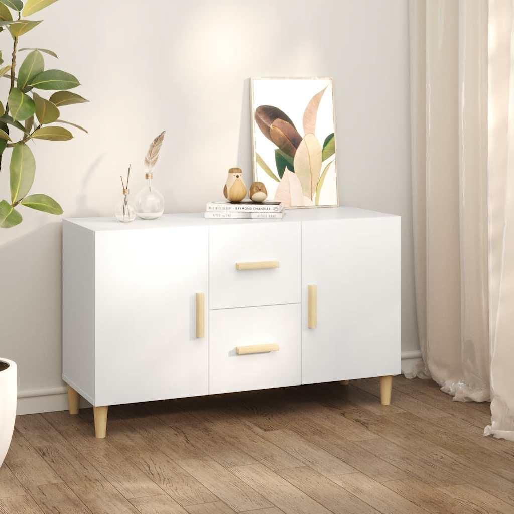 A white sideboard with wooden legs and handles, decorated with a painting and small decorative items, placed on a wooden floor next to a plant and curtains.