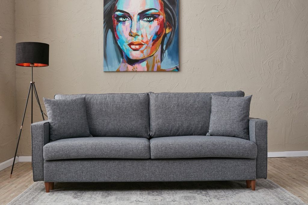 A grey sofa is shown in a living room setting with a painting on the wall and a floor lamp.