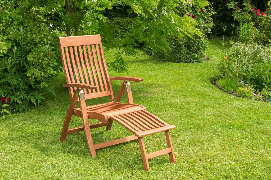 A wooden deck chair with an extended footrest sits on a lush green lawn in a garden setting.