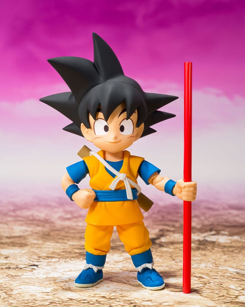 A Son Goku action figure is standing on a sandy surface with a pink sky in the background.
