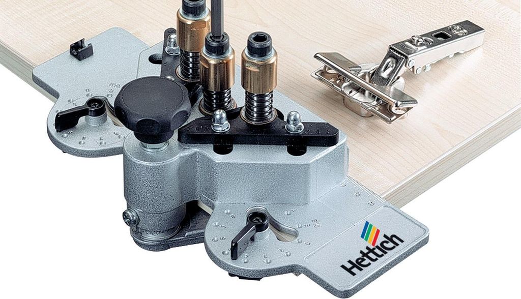 A Hettich jig for drilling holes for hinges is shown on a wooden surface, with a hinge placed next to it.