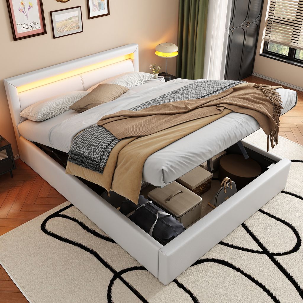 A white upholstered bed with storage is shown with the storage compartment open, revealing items inside. The bed is made up with bedding and blankets.