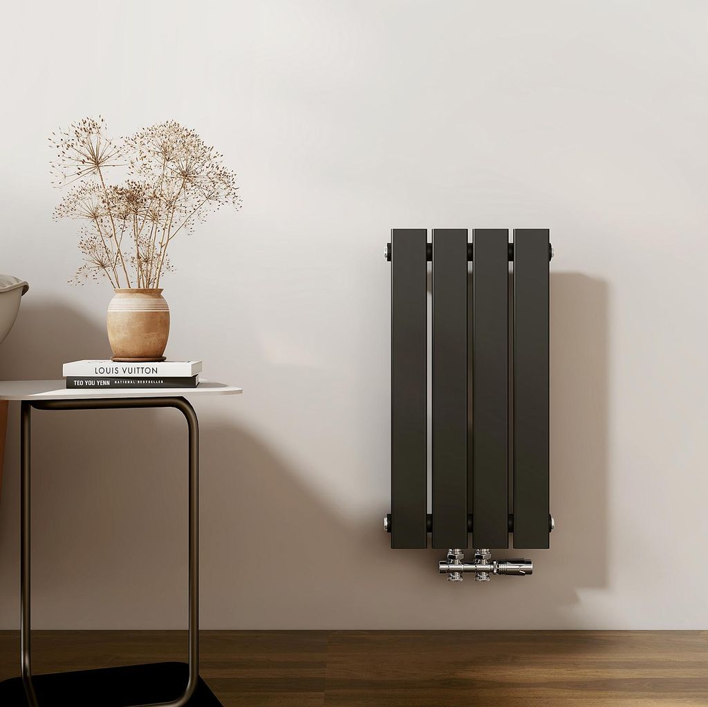 A vertical panel radiator mounted on a light gray wall, next to a small side table with books and a vase of dried flowers.