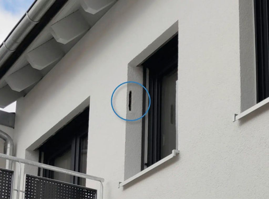 A close-up view of a white building exterior, showing a section of the wall with a window and a small, dark rectangular element highlighted by a blue circle.