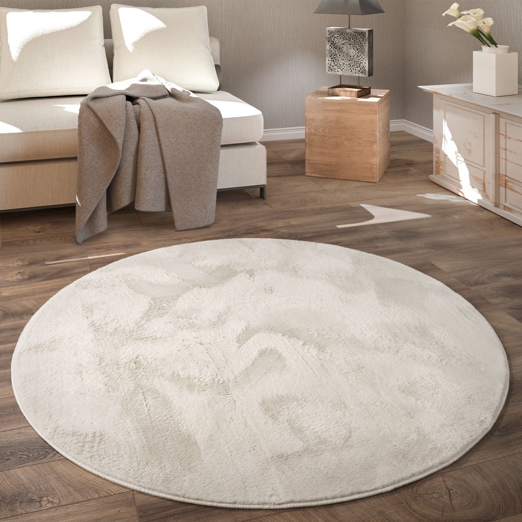 A round, fluffy rug is placed on a wooden floor in a living room setting, with a sofa and side table in the background.