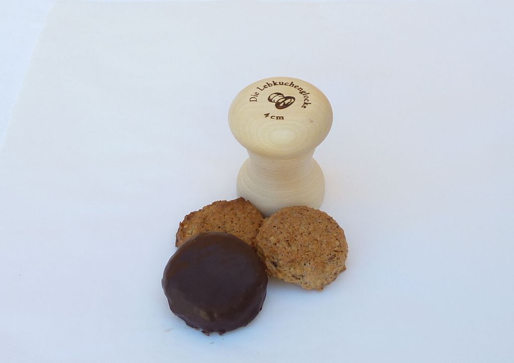 A wooden Lebkuchenglocke stamp with three cookies in front of it on a white background.