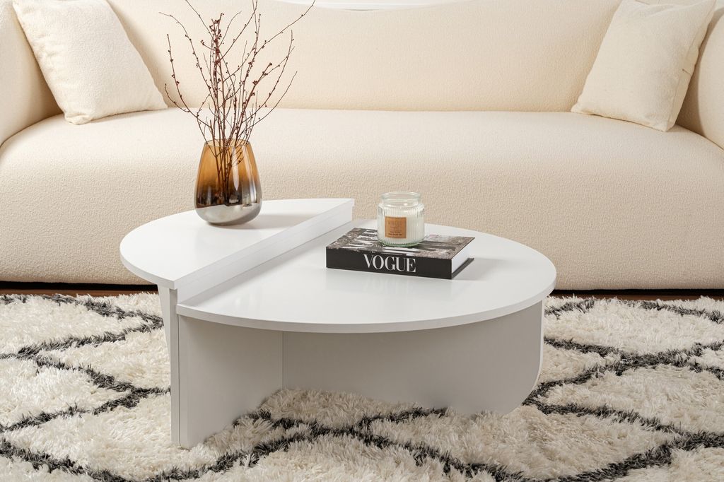 A white coffee table with a vase of branches and a book on it, placed on a patterned rug in front of a cream-colored sofa.
