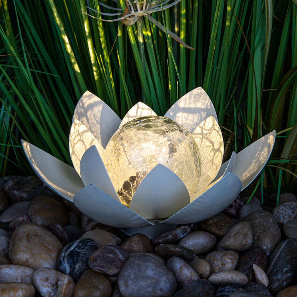A solar-powered lotus flower garden light sits on a bed of river stones, surrounded by lush green foliage.
