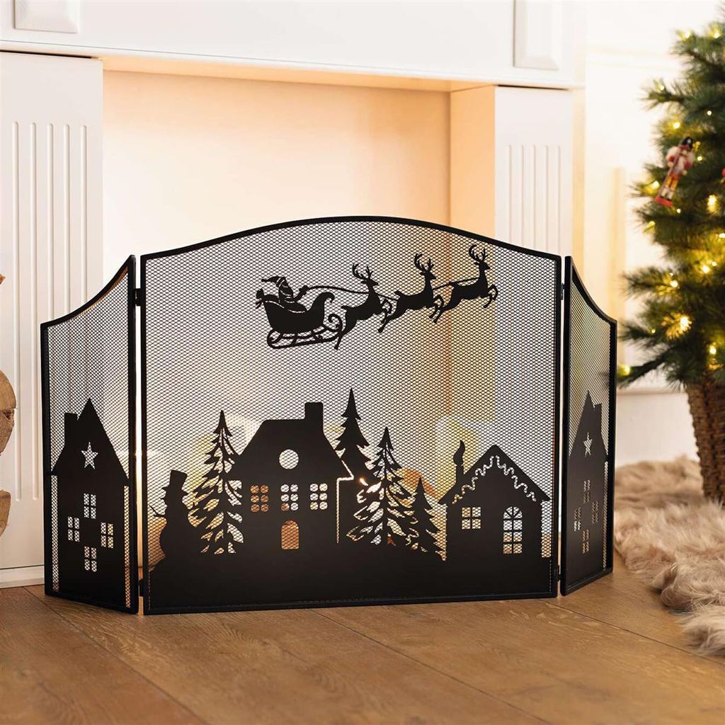 A black metal fireplace screen with a Christmas scene silhouette is placed in front of a fireplace, with a decorated Christmas tree visible to the right.