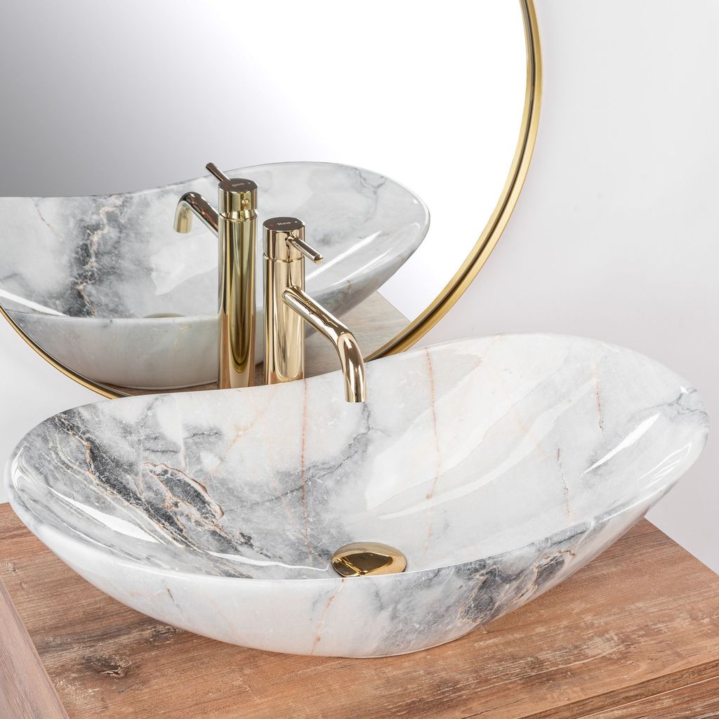 A marble basin with gold faucets is shown on a wooden surface, with a round mirror in the background.
