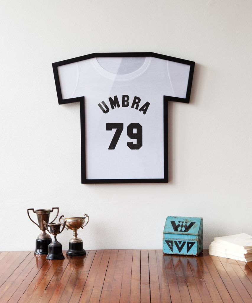 A framed t-shirt artwork hangs on a white wall above a wooden surface with trophies and a decorative box.