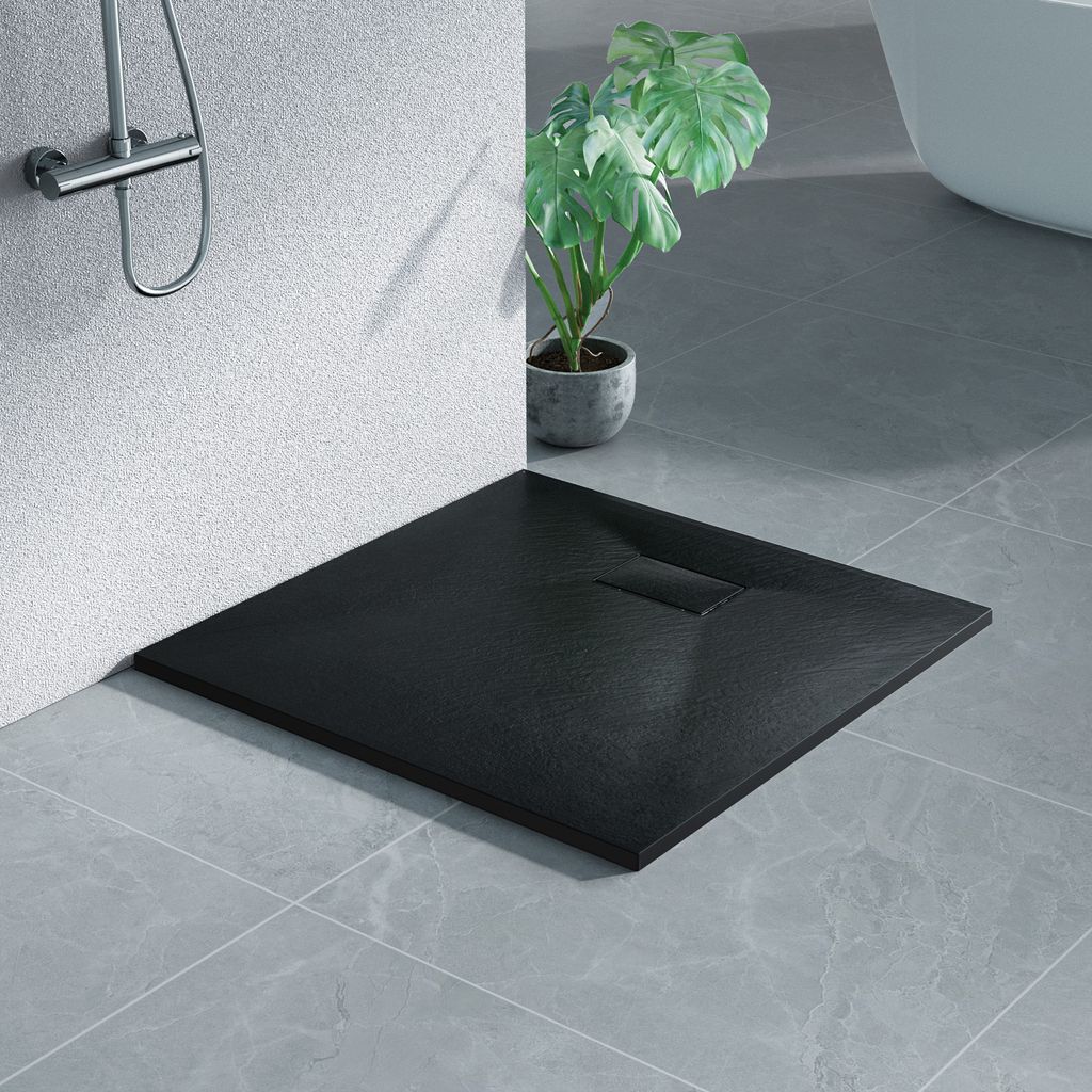A black square shower tray with a stone-like texture is placed on a tiled floor in a bathroom setting. A shower head is visible on the wall, and a potted plant is in the background.