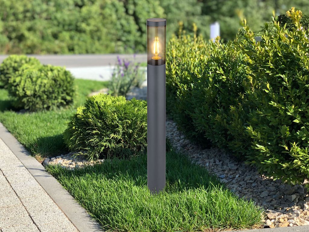 A modern outdoor lamp post stands in a garden setting with manicured bushes and grass.